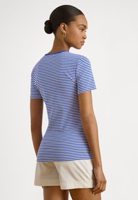 Žena s vlasmi zviazanými do drdolu, nosí zlaté kruhové náušnice, modro-biele pruhované tričko a béžové šortky, smeruje doprava na bielom pozadí.