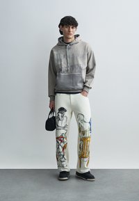 Γκρι φούτερ με κουκούλα και κείμενο, συνδυασμένο με λευκό παντελόνι με πολύχρωμα καλλιτεχνικά σχέδια. Κρατάει μικρή μαύρη τσάντα, φοράει μαύρα παπούτσια.