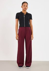 Jonge vrouw die in een ontspannen houding staat, gekleed in een zwart shirt met korte mouwen en rits, en brede bordeauxrode broek met zwarte schoenen.