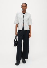 Γκρι πλεκτή ζακέτα με κοντά μανίκια, κλείσιμο με κορδόνι μπροστά, φορεμένη πάνω από μαύρη μπλούζα και μαύρο παντελόνι με φαρδιά πόδια, συνδυασμένη με μαύρη τσάντα χειρός.