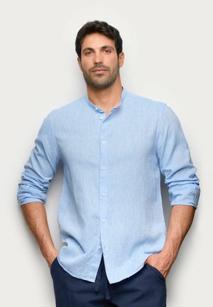 Uomo con capelli scuri e barba, che indossa una camicia azzurro chiaro con bottoni e maniche arrotolate e pantaloni scuri, in piedi con le mani nelle tasche.