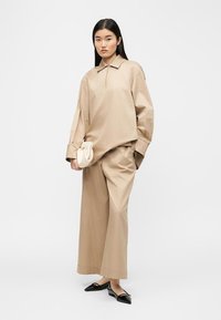 Γυναίκα που φοράει φαρδιά μπεζ πουκάμισο και παντελόνι με φαρδιά πόδια, κρατώντας μια μικρή λευκή τσάντα χειρός και μαύρες μυτερές μπαλαρίνες, στέκεται μπροστά σε λευκό φόντο.