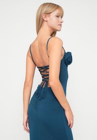 Τιρκουάζ φόρεμα με εφαρμοστή σχεδίαση, με λεπτές τιράντες και δέσιμο στην πλάτη. Διαθέτει ραμμένες υφές και μια φουντωτή λεπτομέρεια στο μπροστινό μέρος.