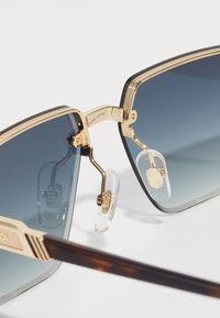 Lunettes de soleil avec une monture carrée, décoration en or, verres dégradés passant du bleu foncé au bleu clair, et plaquettes de nez transparentes ; branches au motif écaille.
