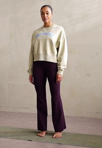 Beige cropped sweatshirt with blue text, paired with fitted burgundy flared leggings. Model stands barefoot on a light green mat.
