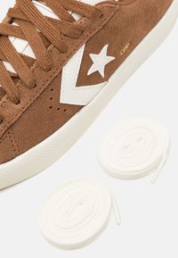 Bruna mockaskor med vita accenter, med en stjärnlogotyp, runda skosnören och en strukturerad gummisula. Två vita skosnören ligger bredvid skon.