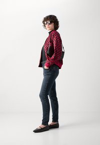 Červená kostkovaná bomber bunda, černý top, tmavé skinny džíny a černé ploché boty. Doplňky zahrnují sluneční brýle a malou černou kabelku.
