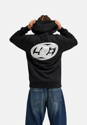 Osoba odwrócona tyłem, trzyma czarną bluzę z dużym białym logo DC Shoes, ubrana w niebieskie dżinsy z takim samym logo na tylnej kieszeni.