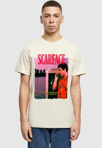 Ungdommelig mann med fregner iført kremfarget t-skjorte med en scene fra "Scarface"-filmen, rød tekst og sitatet "Jeg sier alltid sannheten. Selv når jeg lyver."