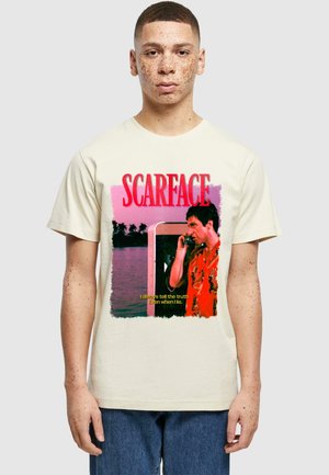 Ungdommelig mann med fregner iført kremfarget t-skjorte med en scene fra "Scarface"-filmen, rød tekst og sitatet "Jeg sier alltid sannheten. Selv når jeg lyver."