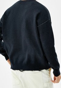 Persona con un suéter negro de dobladillo y puños desgastados, combinado con pantalones blanco roto, visto desde la parte posterior.