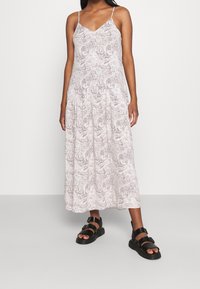 Robe légère sans manches avec une silhouette fluide, ornée d'un motif paisley discret dans des tons beiges et crème. Portée avec des sandales noires.