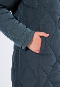 Giacca puffer trapuntata di un verde bluastro scuro con un motivo a diamante, dotata di una tasca laterale e di un tessuto testurizzato, leggermente lucido.