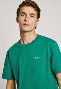 Green cotton t-shirt with short sleeves and a crew neckline. Features a white logo on the left chest area. Smooth texture.