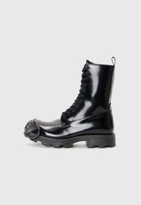Musta lakkapintainen taistelukenkä kiiltävällä pinnalla, nauhakiinnitys, paksu kumipohja ja erottuva etuosan metalliosuus.