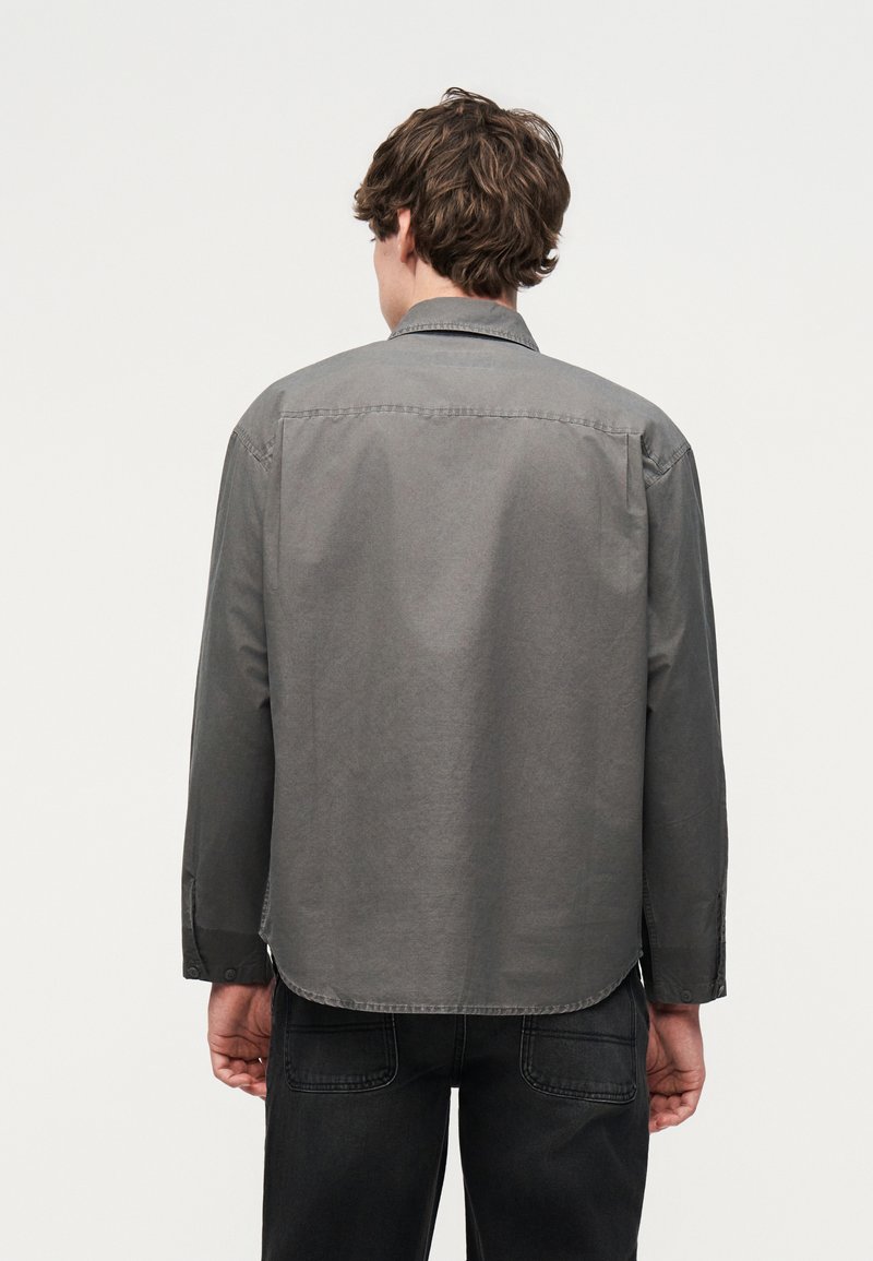 Uomo con capelli corti castani, indossa una camicia grigia a maniche lunghe abbottonata e pantaloni neri, visto di spalle su uno sfondo semplice.