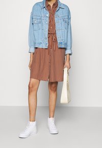 Veste en jean sur une robe chemise à rayures marron et blanches, associée à des baskets montantes blanches et à un sac à main texturé de couleur crème.