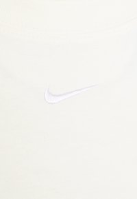 Un tessuto di colore crema con una texture morbida presenta un piccolo logo Swoosh Nike ricamato in bianco sul lato sinistro.