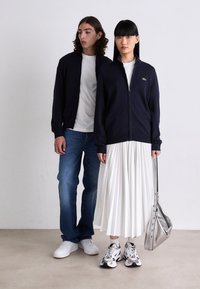 Due modelli stanno in piedi contro uno sfondo semplice indossando maglioni a zip blu navy; uno abbinato a jeans e sneakers bianche, l'altro con una gonna plissettata bianca e una borsa argentata.
