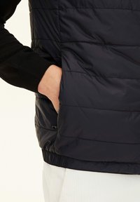 Svart pufferväst med horisontell sömnad, som har en sidoficka och en elastisk fåll. Ytmaterialet verkar blankt och slätt.