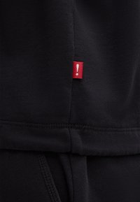 Felpa nera con un piccolo etichetta rossa con un punto esclamativo sul lato, realizzata in morbido cotone con una texture liscia.