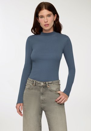 Maglione a maniche lunghe a collo alto in tessuto blu spento a costine, abbinato a jeans grigi sbiaditi a vita alta con chiusura a bottone e una sottile texture.