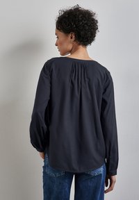 Marineblaues Langarmshirt mit lockerer Passform, rundem Halsausschnitt und gerafftem Detail auf der Rückseite. Getragen mit blauen Jeans.