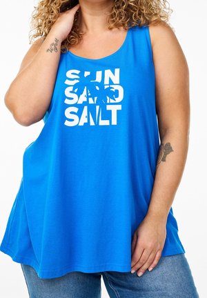 Krøllet person iført en blå tanktop med "SUN SAND SALT" og en palme grafik, kombineret med blå jeans.