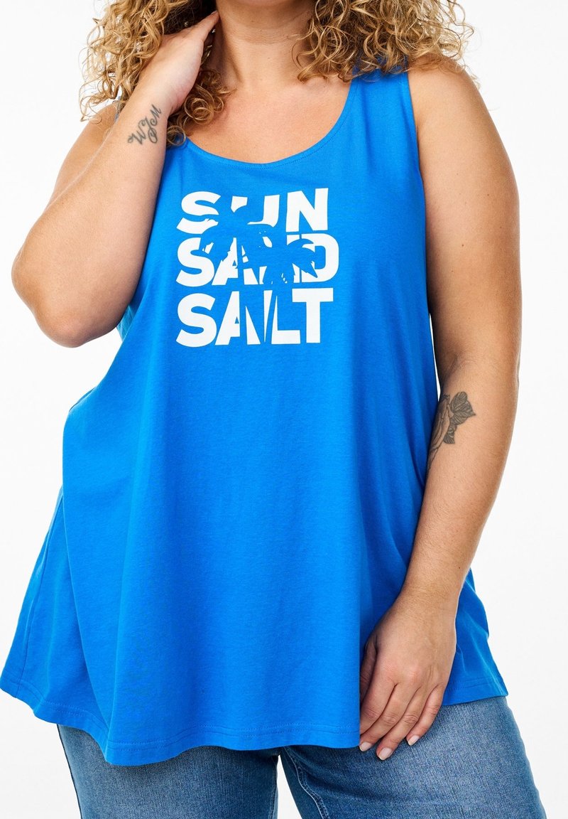 Personne aux cheveux bouclés portant un débardeur bleu avec « SUN SAND SALT » et un motif de palmier, associé à un jean bleu.