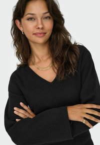 Žena s dlouhými vlnitými vlasy, oblečená v černém svetr s výstřihem do V, se zlatými řetízky, náušnicemi a prsteny, se zkříženýma rukama, mírně se usmívající.
