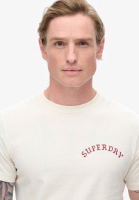 Camiseta de manga corta color crema confeccionada en algodón, con un logo rojo de "SUPERDRY" curvado en la parte izquierda del pecho. Textura suave en toda la prenda.