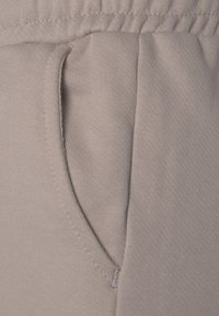 Gros plan sur un pantalon de survêtement en tissu beige montrant une ceinture élastique et une poche latérale avec des détails de couture visibles.