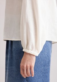Blusa bianca in lino con maniche a sbuffo, caratterizzata da polsini arricciati. Abbinata a pantaloni in denim blu testurizzati, aggiunge contrasto e interesse visivo.