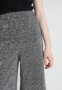 Grå vida byxor med en texturerad tygkvalitet som har vertikala ribbor och utspridda silverpaljetter. Synlig handdetalj till höger.