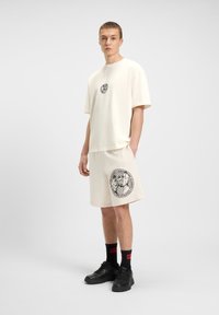 Creme T-shirt med rund hals og sort grafisk logo, parret med matchende shorts med stort sort print. Sorte sneakers og sokker med røde accenter.