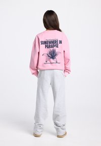 Roze sweatshirt met de tekst "SOMEWHERE IN PARADISE" en een bloemenafbeelding op de achterkant; gecombineerd met grijze joggingbroek en beige schoenen.