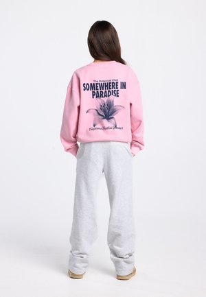 Pink sweatshirt with "SOMEWHERE IN PARADISE" text and floral graphic on the back; paired with gray sweatpants and tan shoes.