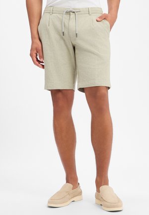 Beige knielange Shorts mit Kordelzug, getragen von einer Person mit teilweise in den Taschen steckenden Händen, kombiniert mit hellbeigen Slippern.