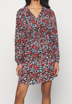 Kobieta ubrana w czarną sukienkę w kwiaty w kolorach czerwonym, różowym i niebieskim, z długimi rękawami, dekoltem w serek i marszczoną talią.