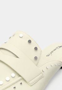 Chaussure blanche cassée à enfiler avec des clous ronds argentés et une bride sur le pied, montrant des coutures détaillées et une partie du logo de la semelle intérieure.