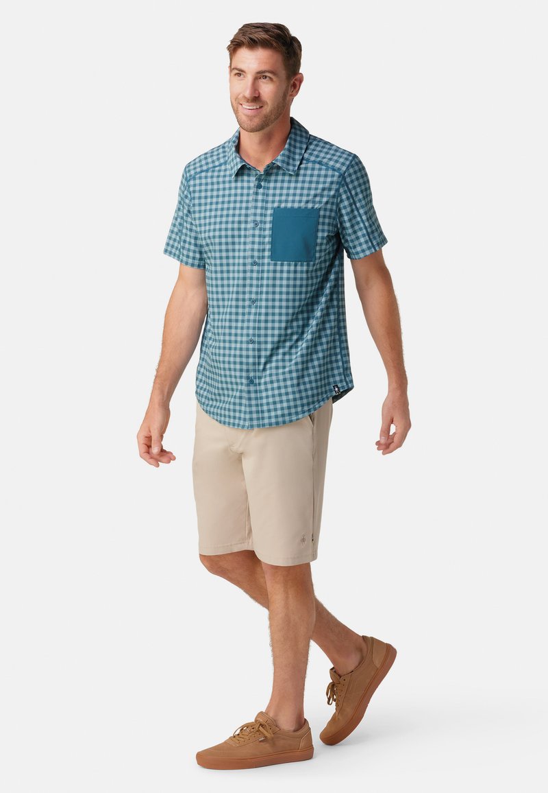 Kortærmet, ternet skjorte i teal og hvid med en ensfarvet teal lomme. Kombinationen med beige shorts og brune sneakers.