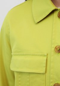 Limonkowo-zielona, teksturowana kurtka koszulowa z ostrym kołnierzykiem, kieszenią na piersi i brązowymi guzikami, z kontrastującymi detalami przeszycia.