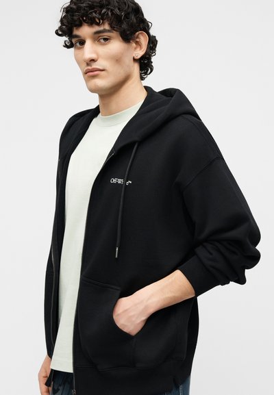 Νεαρός με σγουρά σκούρα μαλλιά, φορώντας μαύρη ζακέτα με φερμουάρ πάνω από λευκό πουκάμισο, με το χέρι στην τσέπη, στραμμένος ελαφρώς προς τα αριστερά.