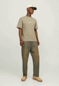 Hombre con gorra beige, camiseta verde oliva, jeans holgados deslavados y zapatos beige de pie contra un fondo claro y liso.