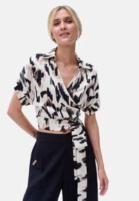 Beżowa bluzka w abstrakcyjny czarno-różowy wzór, z krótkimi rękawami i wiązaniem z przodu, zestawiona z czarnymi spodniami o wysokim stanie.