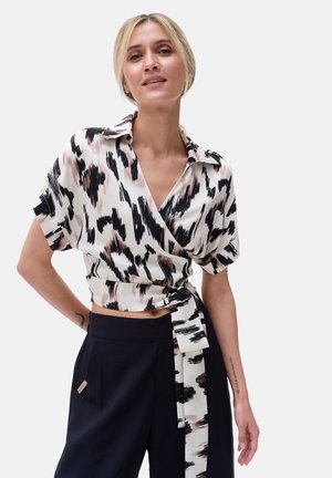 Wrap blouse in beige with black and pink abstract pattern, short sleeves, and front tie detail, paired with black high-waisted pants.