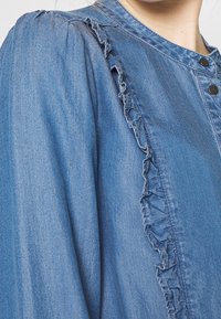 Light blue denim shirt with a ruffled placket, button closure, and gathered shoulder seams, featuring a soft texture.