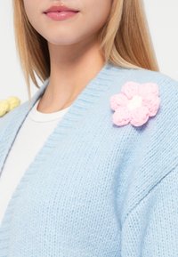 Απαλή, γαλάζια πλεκτή ζακέτα με V-λαιμόκοψη, διακοσμημένη με μια ροζ ανθισμένη απεικόνιση με λευκό κέντρο στον αριστερό ώμο. Απαλή υφή.