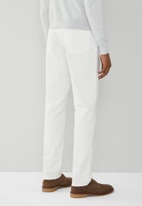 Pantalones de algodón blancos con un corte recto, que cuentan con dos bolsillos traseros y se llevan con zapatos de ante marrones. Suéter gris visible en la parte superior.