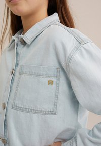 Light blue denim jacket with a button-up front, a chest pocket, and an embroidered "R" detail in gold on the pocket.